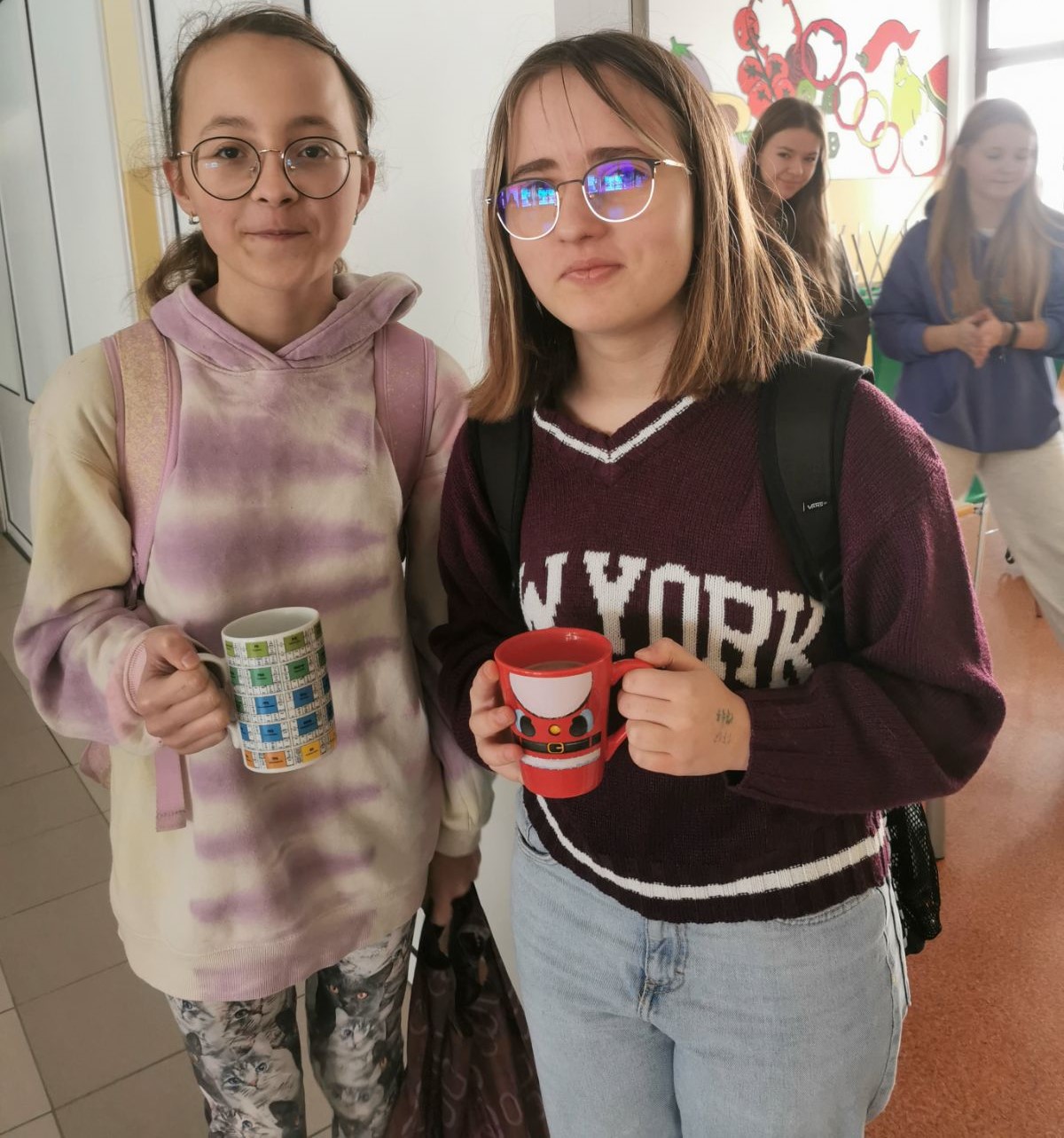 Na tle szkolnej stołówki stoją dwie dziewczynki z kolorowymi kubkami w rękach.
