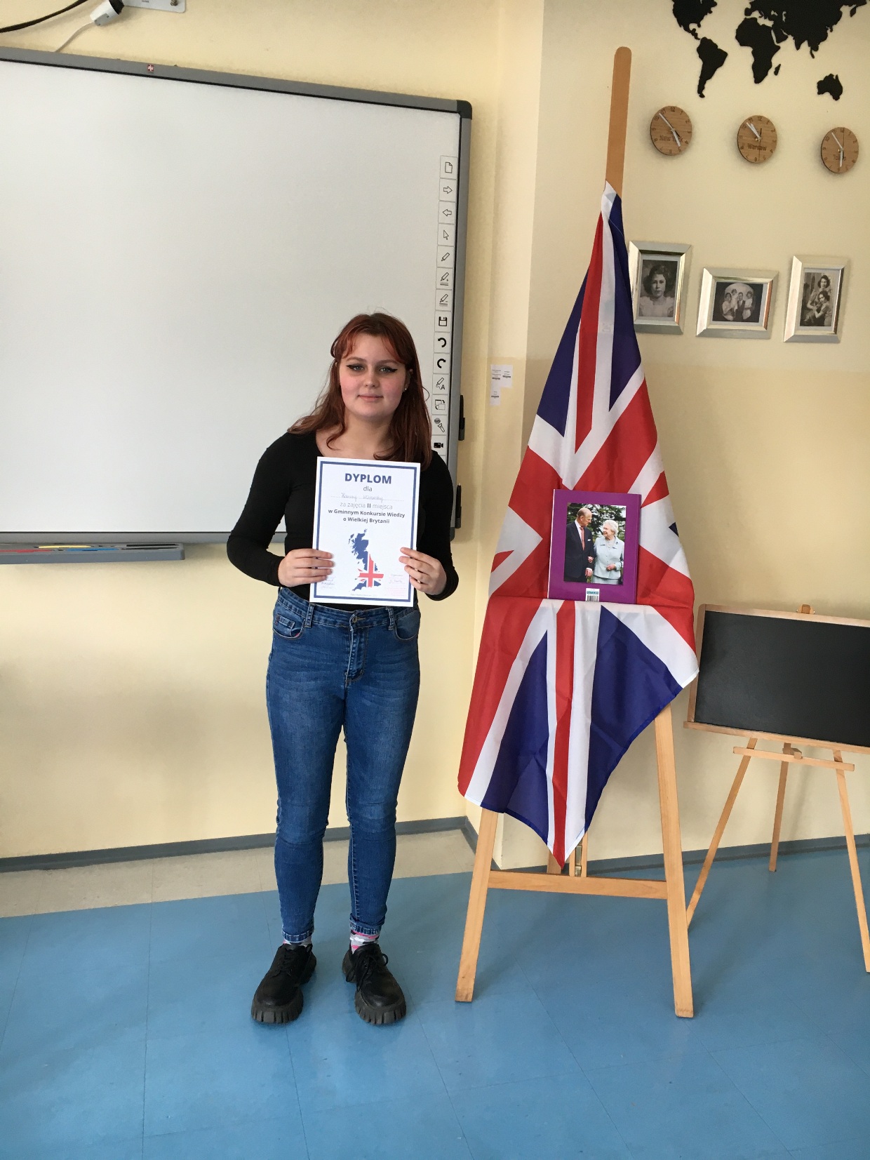 dziewczyna z dyplomem stoi w klasie obok flagi Zjednoczonego Królestwa Wielkiej Brytanii i zdjęcia Królowej Elżbiety i księcia Wilama